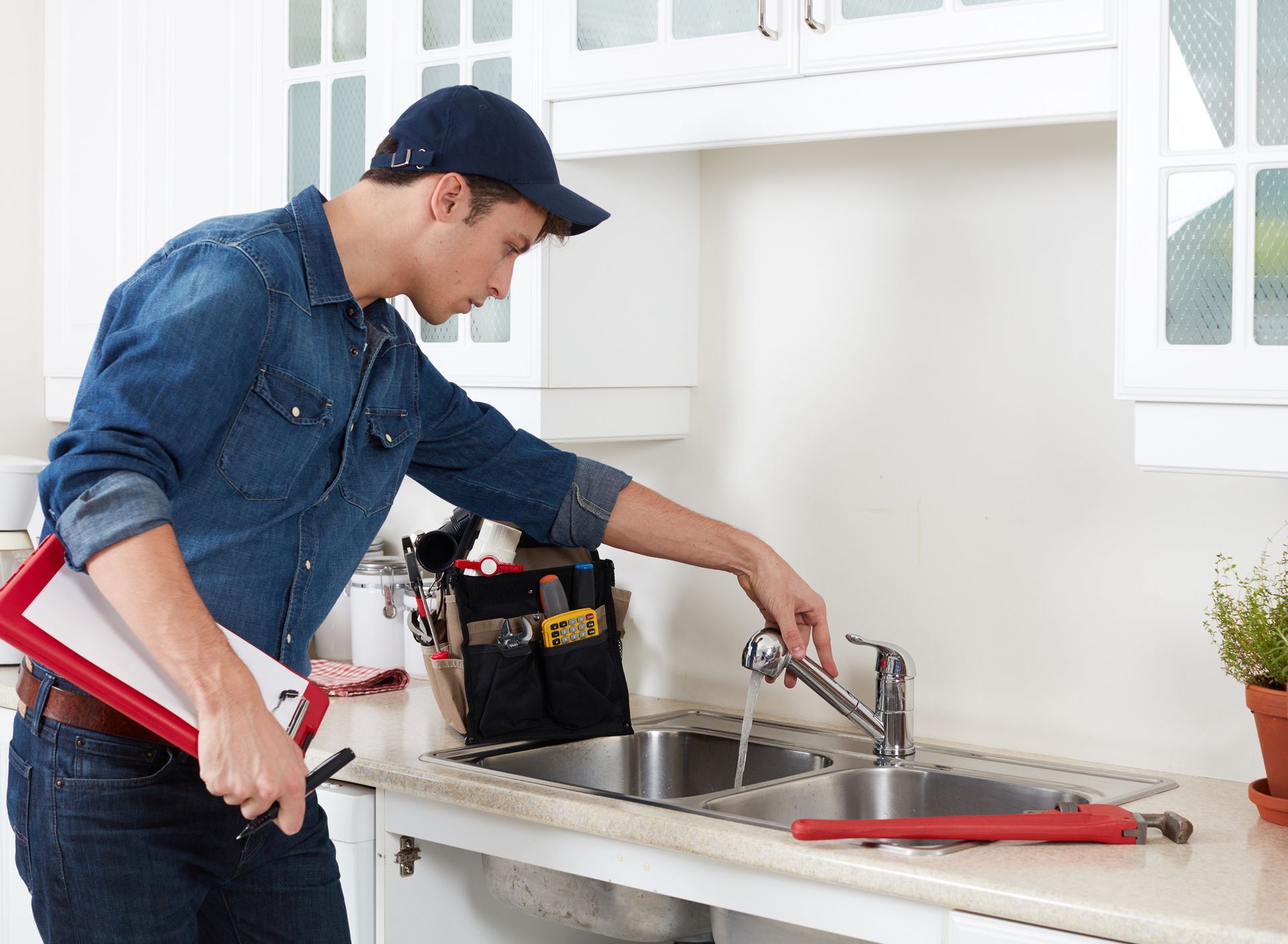 A plumber repairing a kitchen sink faucet with a tool belt and pipe wrench